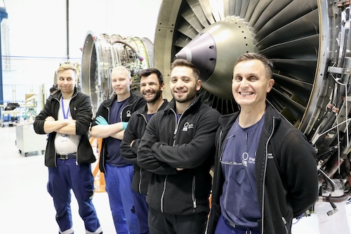 Viisi miestä hymyilee lentomoottorin edessä